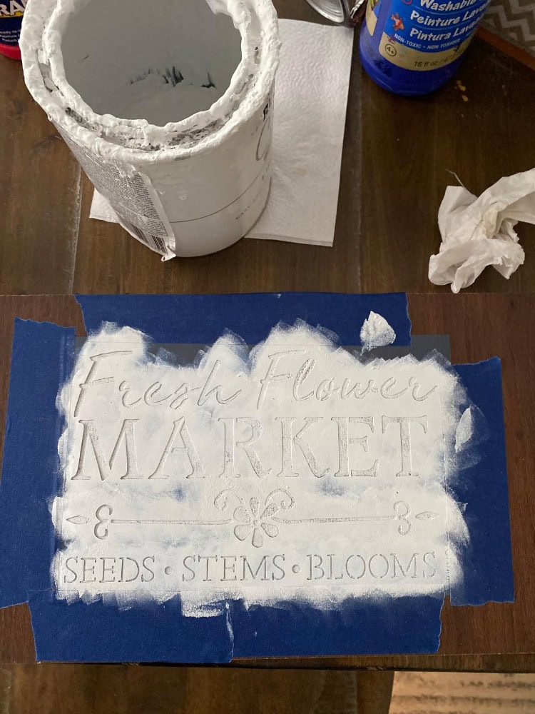 Fresh Flower Market sign stenciled on wood with white paint, featuring seeds, stems, and blooms, surrounded by painting supplies for floral decor and gardening projects.