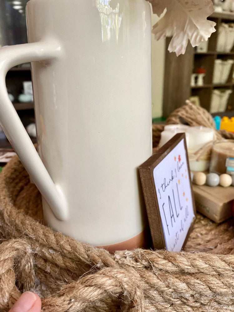 White ceramic mug with handle, part of a cozy home decor display on a rustic jute basket, outdoor-themed accessories and inspirational sign in background, perfect for gift or decorative use.