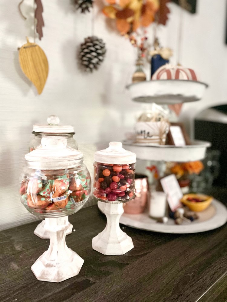 Mini candy jars with colorful sweets on white vintage stands, decorative autumn fall leaves and pinecones on a white wall background, cozy home decor for fall or holiday seasons, A Hundred Affections.