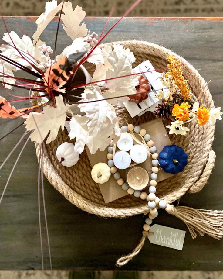 Rustic fall-themed centerpiece with candles, faux pumpkins, seasonal flowers, and a gratitude tag, perfect for autumn decor and Thanksgiving celebrations.
