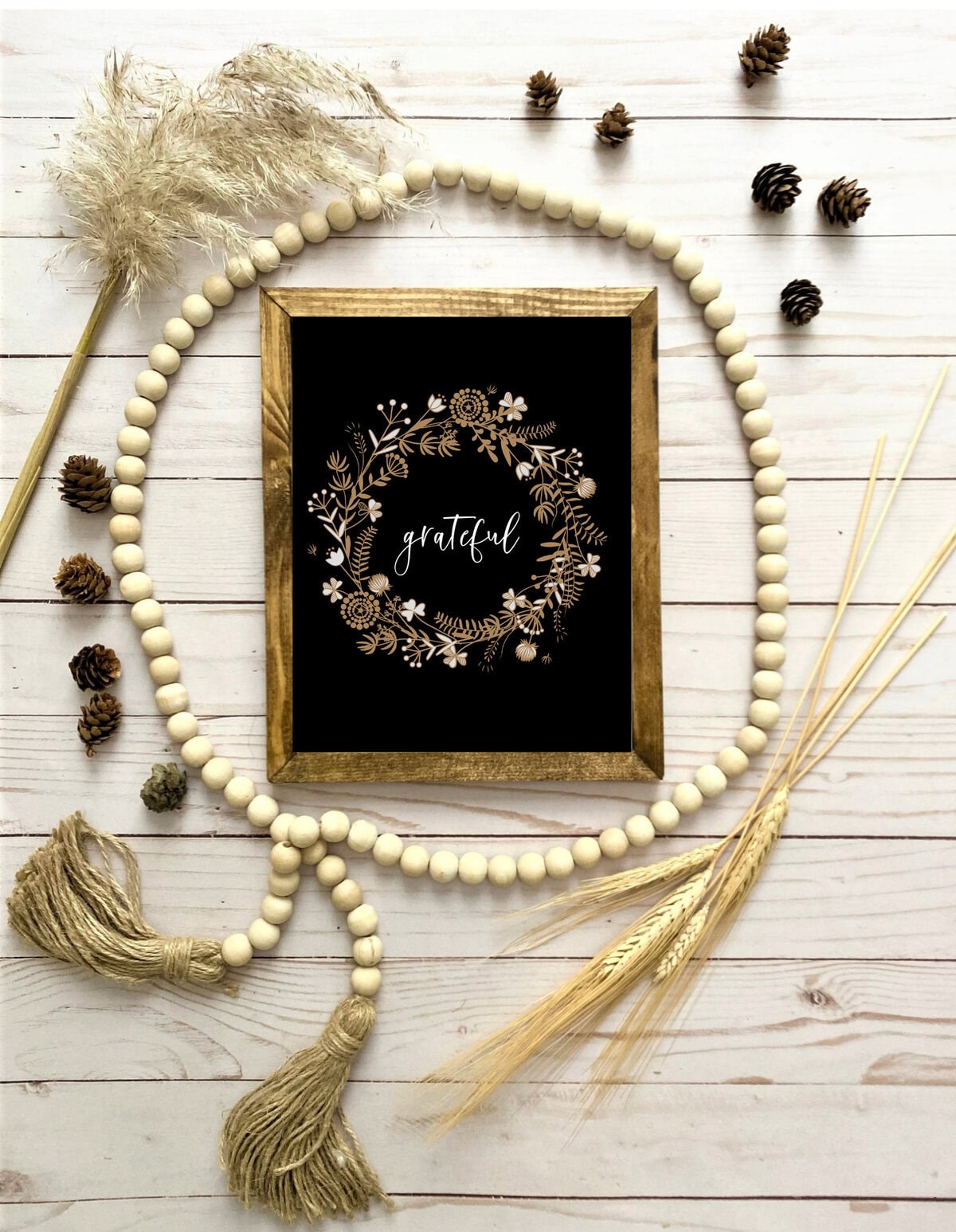 Beaded wood garland, dried pampas grass, pinecones, wheat stalks, and a framed art print with "grateful" in elegant script on a white wooden surface, creating a cozy, rustic, boho-inspired decor setup.