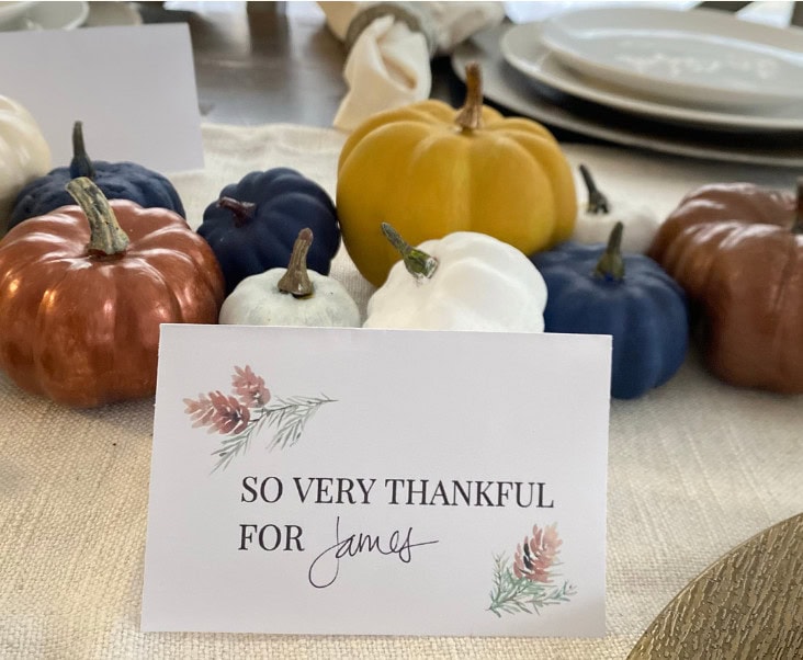 Colorful painted pumpkins in shades of copper, navy, white, gold, and terracotta on a linen tablecloth, with a gratitude card reading "So very thankful for James," perfect for fall or Thanksgiving decor.