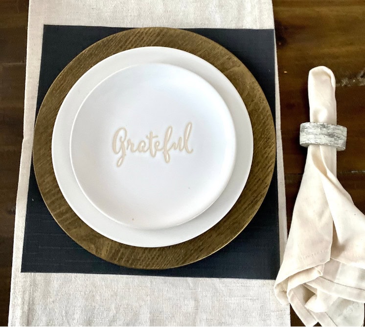 Grateful written on a white plate, rustic wooden charger, minimalist table setting, elegant napkin with napkin ring, mindful gratitude home decor, A Hundred Affections.