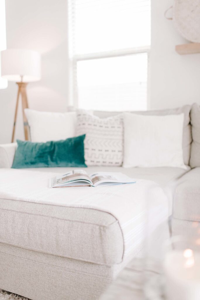 Cozy white living room with a tufted sofa decorated with white and teal throw pillows, an open magazine, modern lamp, and natural light, creating a serene and inviting space for relaxation and home decor inspiration.