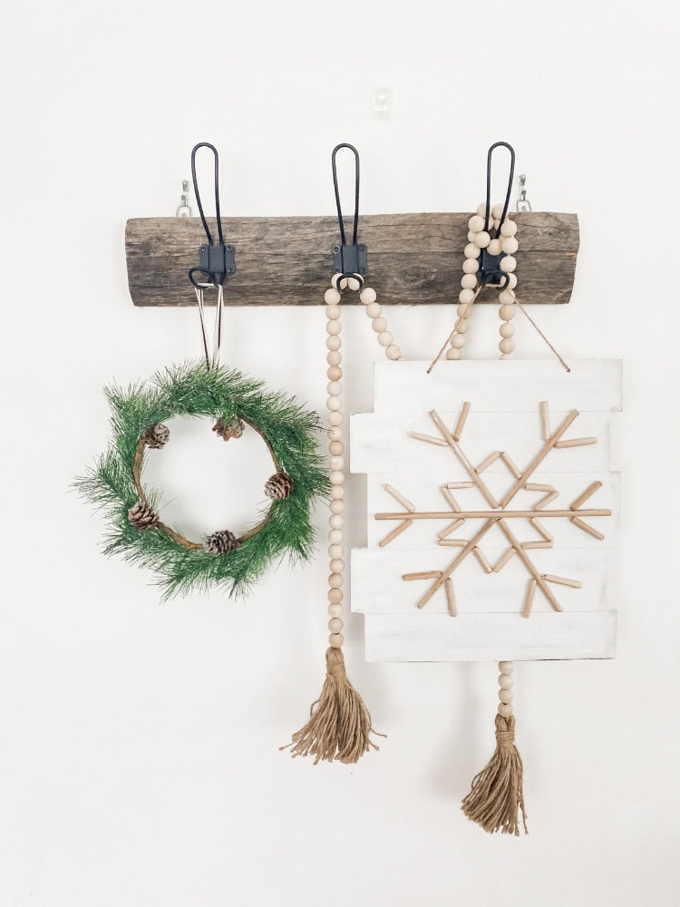 A rustic wooden wall hook display with holiday decor, including a green pine wreath with pinecones, a beaded tassel, and a white wooden snowflake sign with wooden sticks and tassels, perfect for festive holiday home accents.