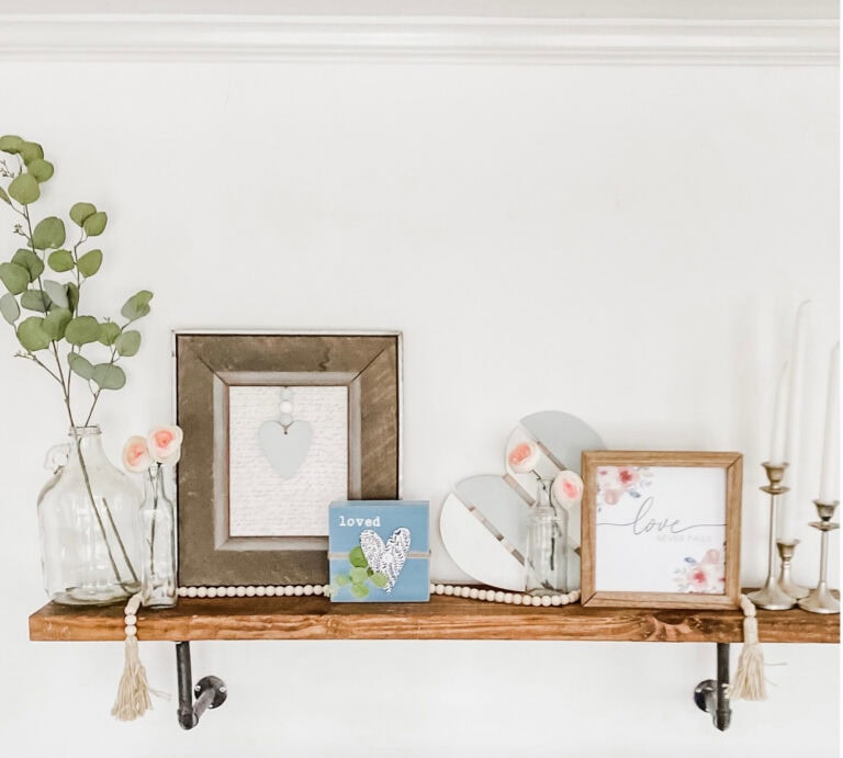 Vintage wooden shelf with decorative signs, glass vases with pink flowers, eucalyptus, and candle holders, creating a charming and romantic home decor setup emphasizing love and affection.