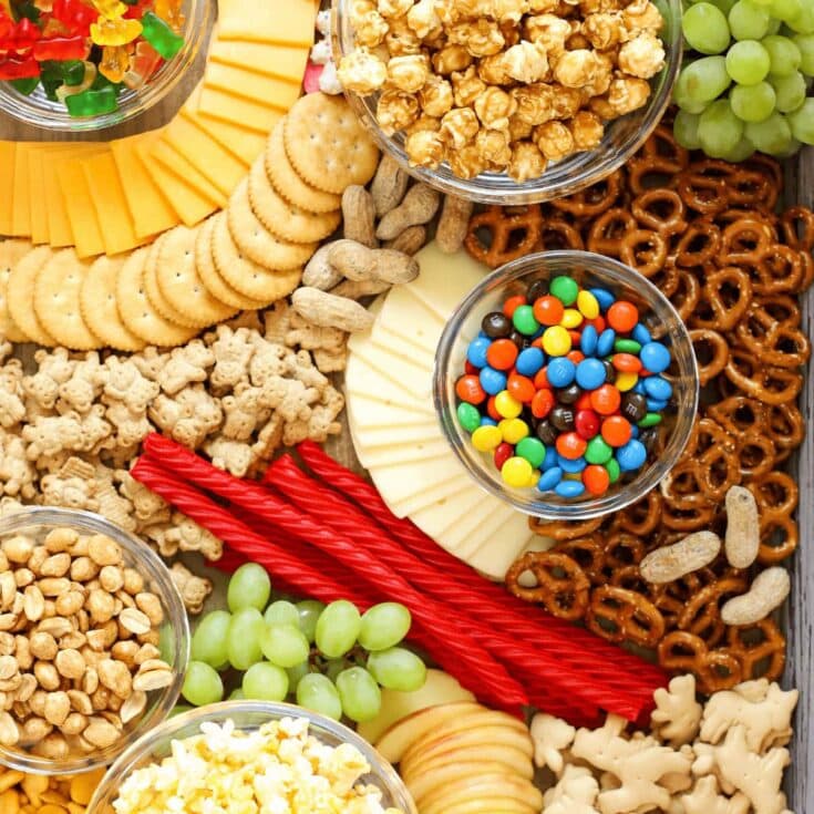 Chewy caramel popcorn, gummy worms, multicolored M&Ms, cheddar cheese slices, pretzels, green grapes, Red Vines, and assorted crackers arranged on a wooden surface for snack platter.