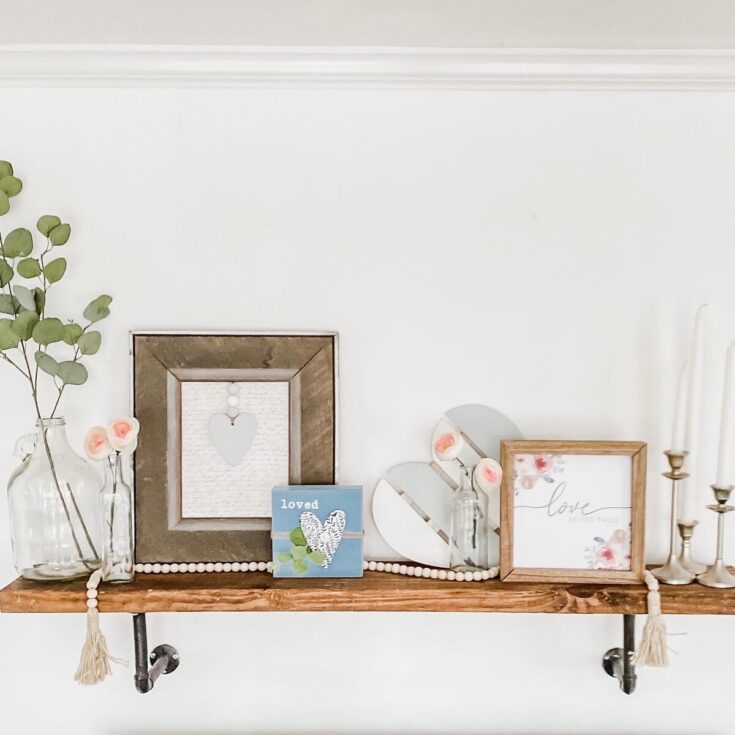 Delicate floral and heart-themed decor on a wooden shelf, featuring framed love quotes, glass vases with pink roses, and candlesticks, perfect for creating a romantic and cozy home ambiance.