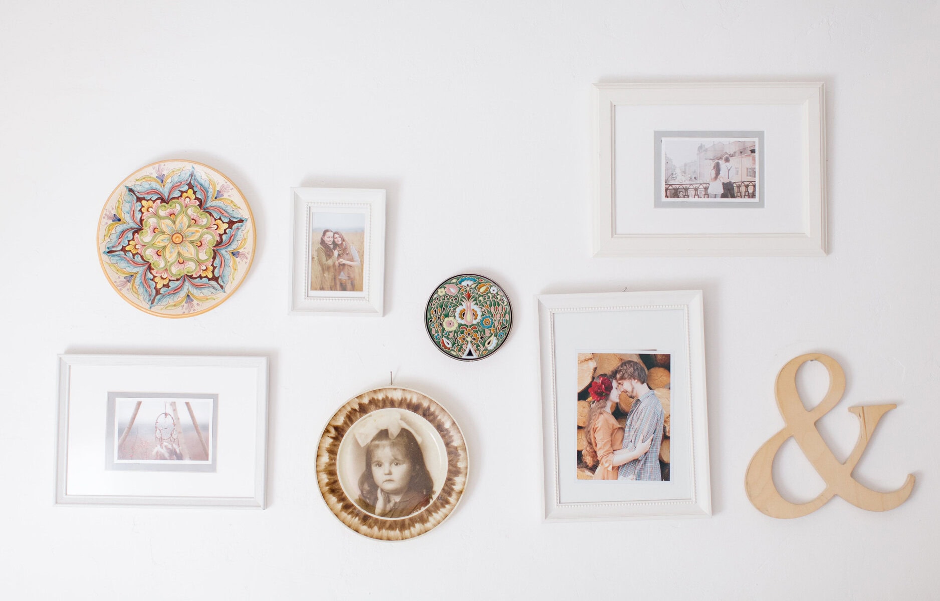 Colorful decorative plates, framed family photos, and an "&" wooden sign displayed on a white wall, creating a charming and personalized home decor arrangement.