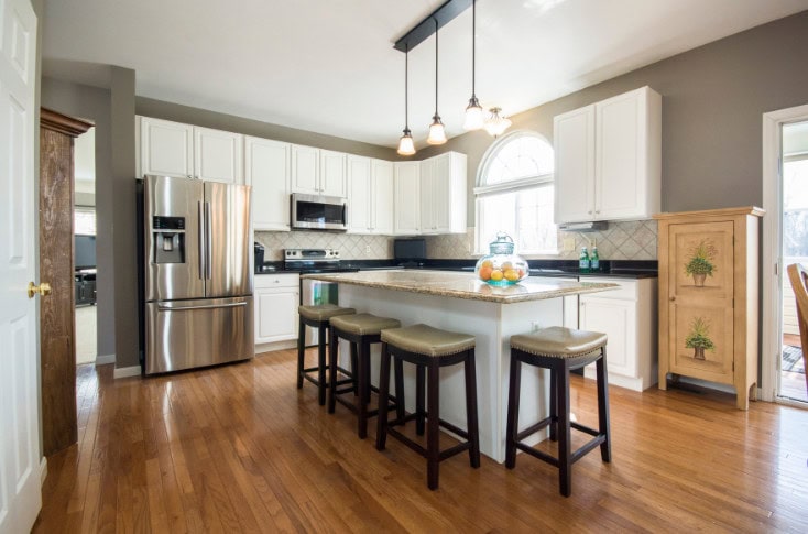 Modern kitchen with white cabinetry, stainless steel appliances, granite countertops, and hardwood flooring, featuring a spacious island with bar stools and ample natural light.