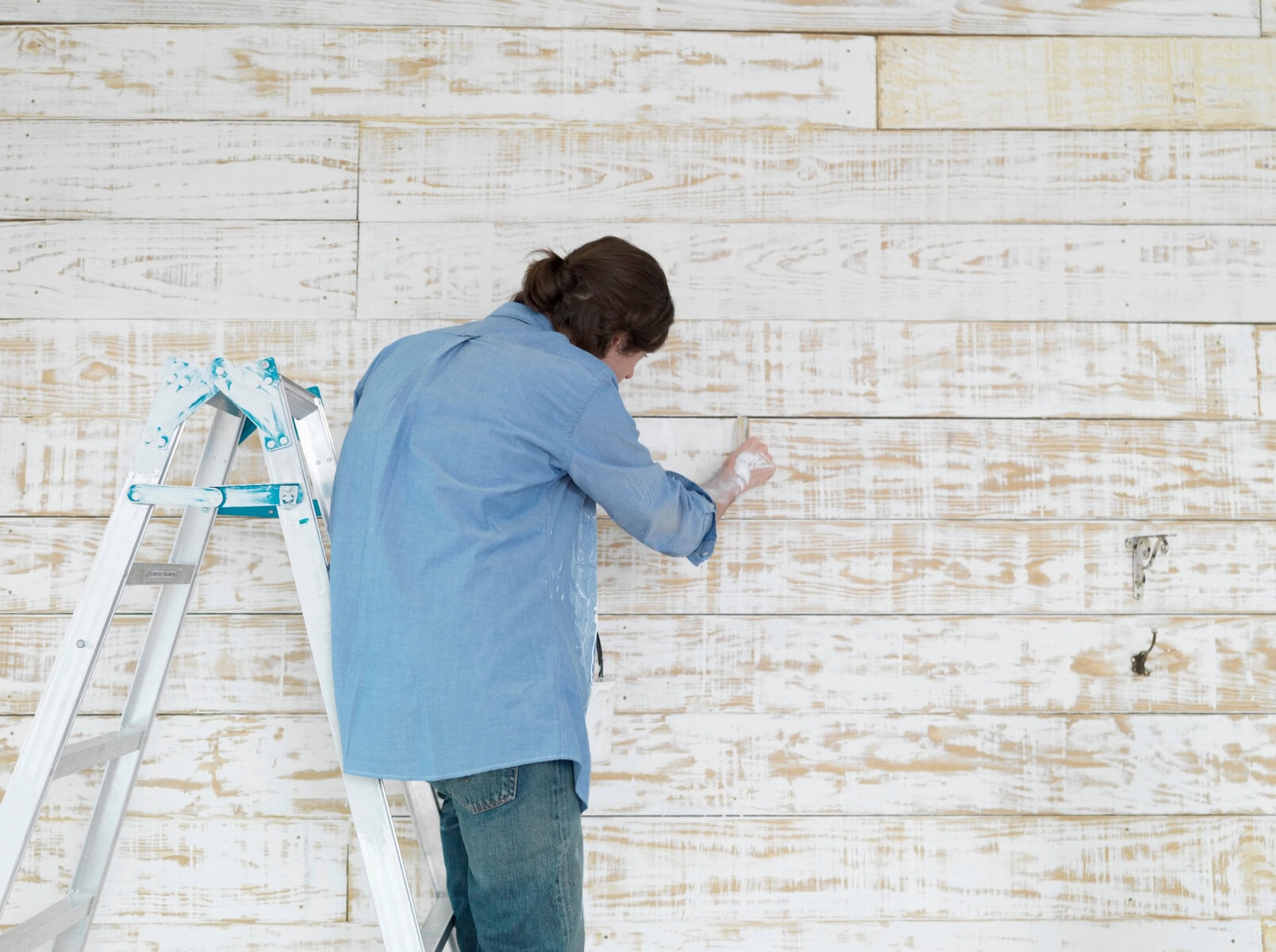 Painted white wooden wall being decorated with a woman painting or distressing, standing on a ladder, for a cozy, rustic home decor look.
