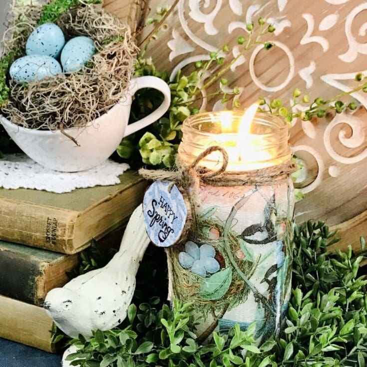 Vintage spring decor with a lit candle in a floral jar, a white ceramic bird, a teacup filled with speckled blue eggs and moss, surrounded by lush greenery and decorative wooden details.