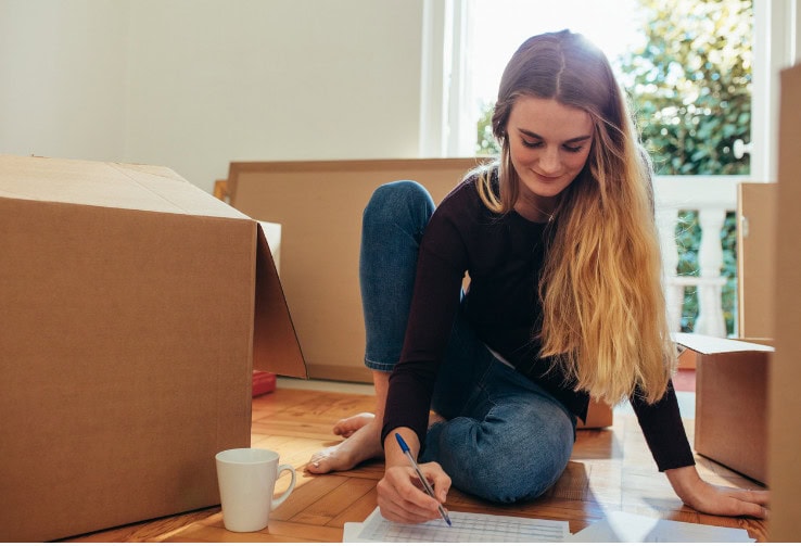 Moving boxes packed for a new home, young woman writing a checklist on the floor, representing moving, organization, and new beginnings, in a bright, cozy home environment.