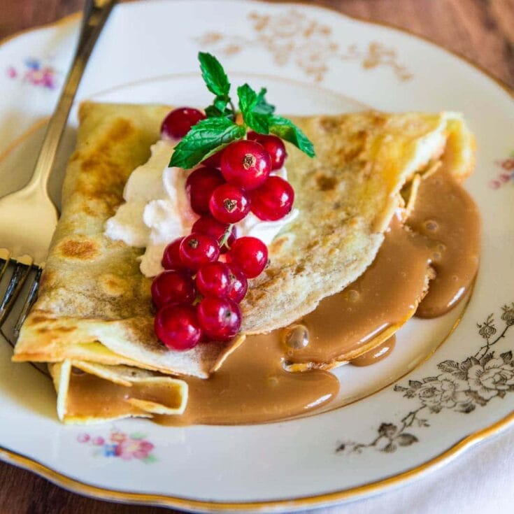 Delicious crepe with caramel sauce, topped with fresh red currants, whipped cream, and mint leaves on a vintage floral plate. Perfect for breakfast or brunch and ideal for using high-quality ingredients.