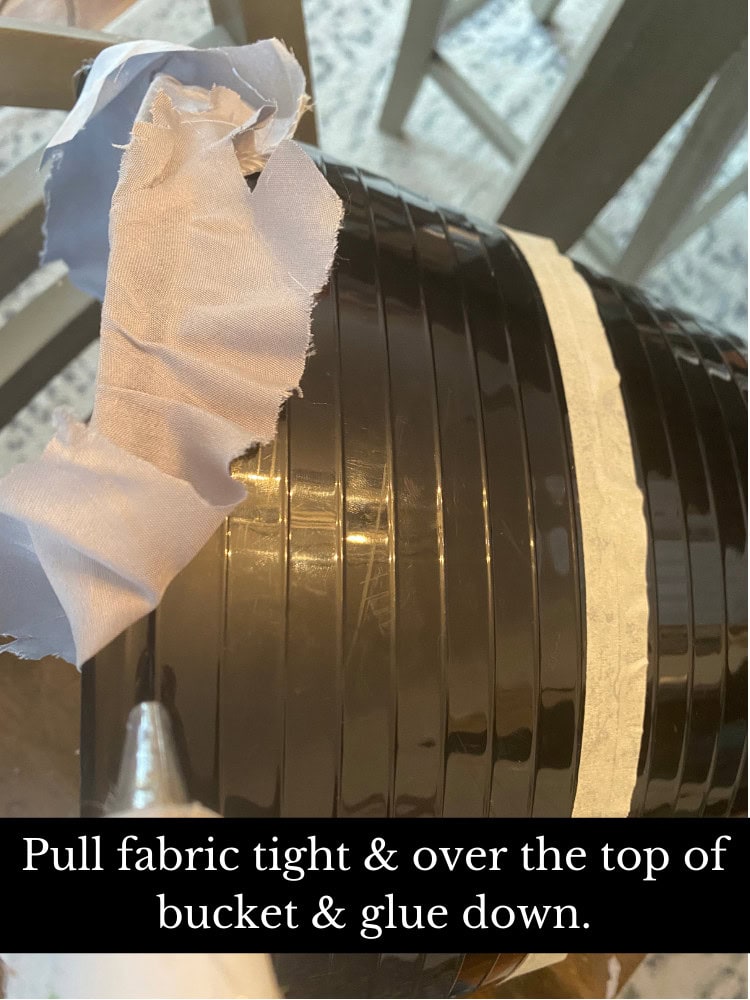 Pulling fabric tight over the top of a black bucket for DIY craft project, using glue to secure the fabric in place.