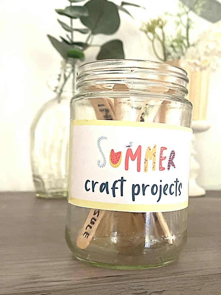 Summer craft projects jar with wooden stir sticks and colorful label on a wooden surface, ideal for DIY summer activities and creative crafts.