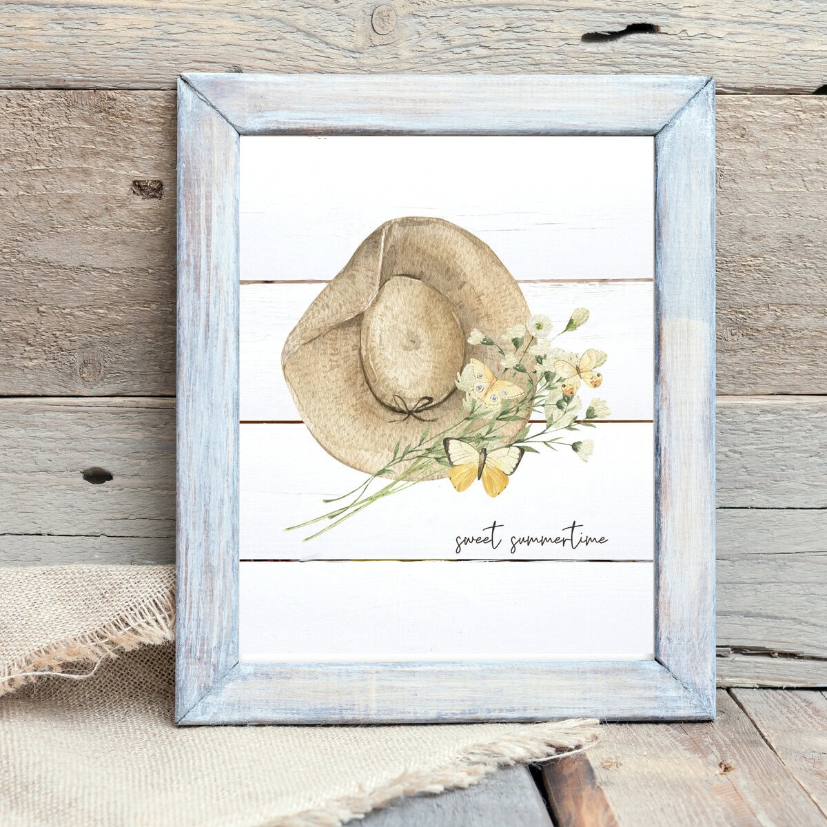 Soft watercolor illustration of a straw hat with delicate flowers and butterflies, featuring the phrase "sweet summertime" on a white wooden background, perfect for summer home decor.