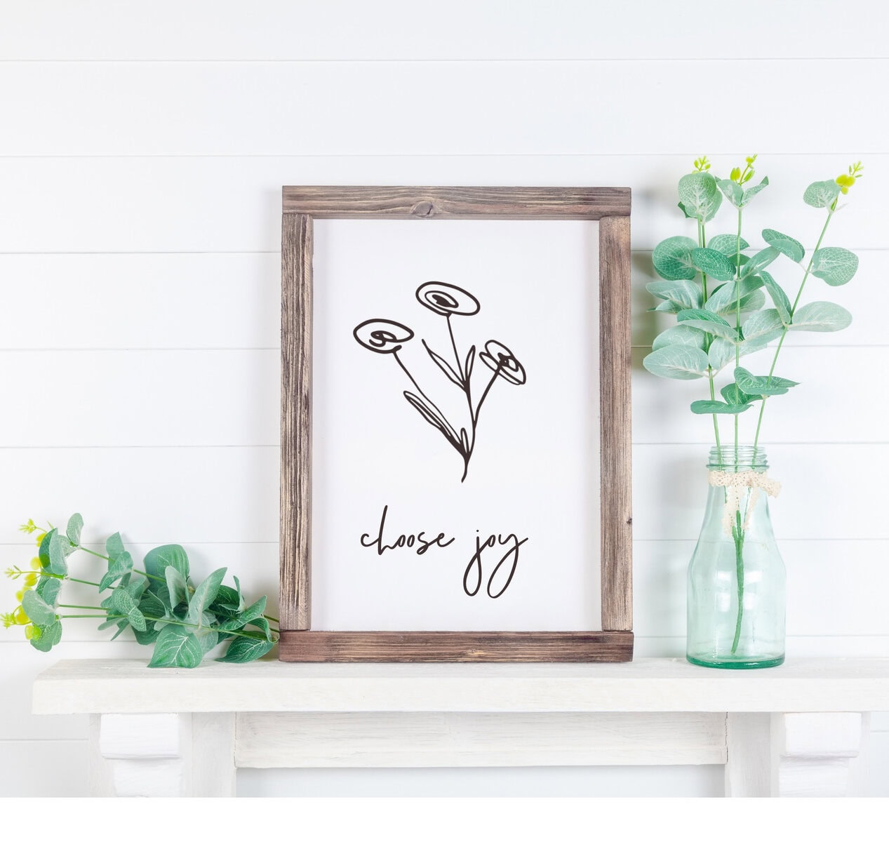 Minimalist wall art with a black line drawing of flowers and the phrase "choose joy" in elegant script, displayed on a wooden frame, surrounded by fresh green plants in glass vases.