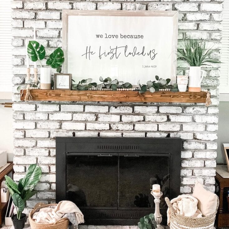 White brick fireplace with wooden mantel decorated with green plants, candles, and a framed quote "we love because He first loved us" from 1 John 4:19 in a cozy home setting.