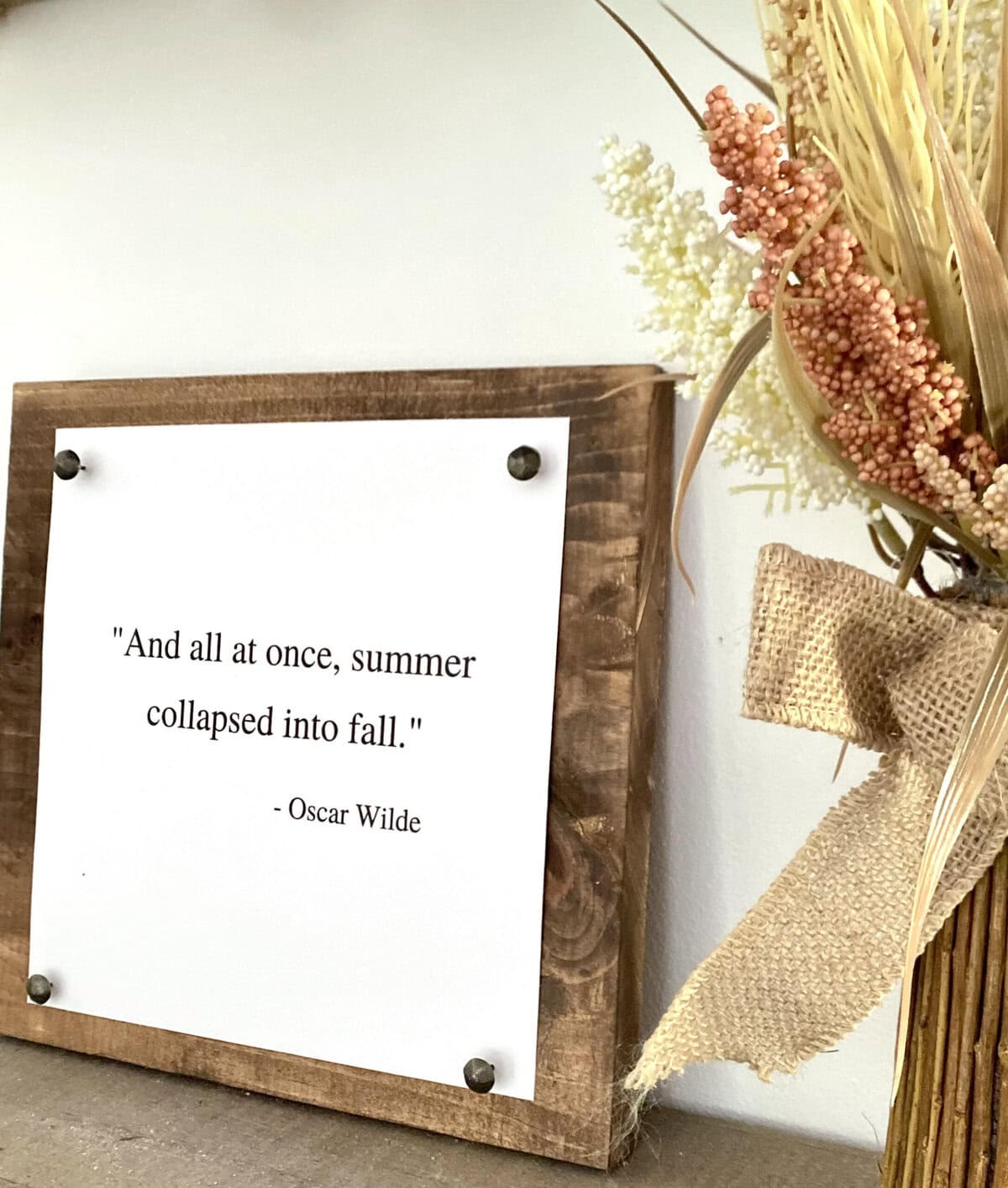 Rustic wooden frame with Oscar Wilde quote on fall transitioning from summer to autumn, decorated with a burlap bow and surrounded by dried floral arrangements.