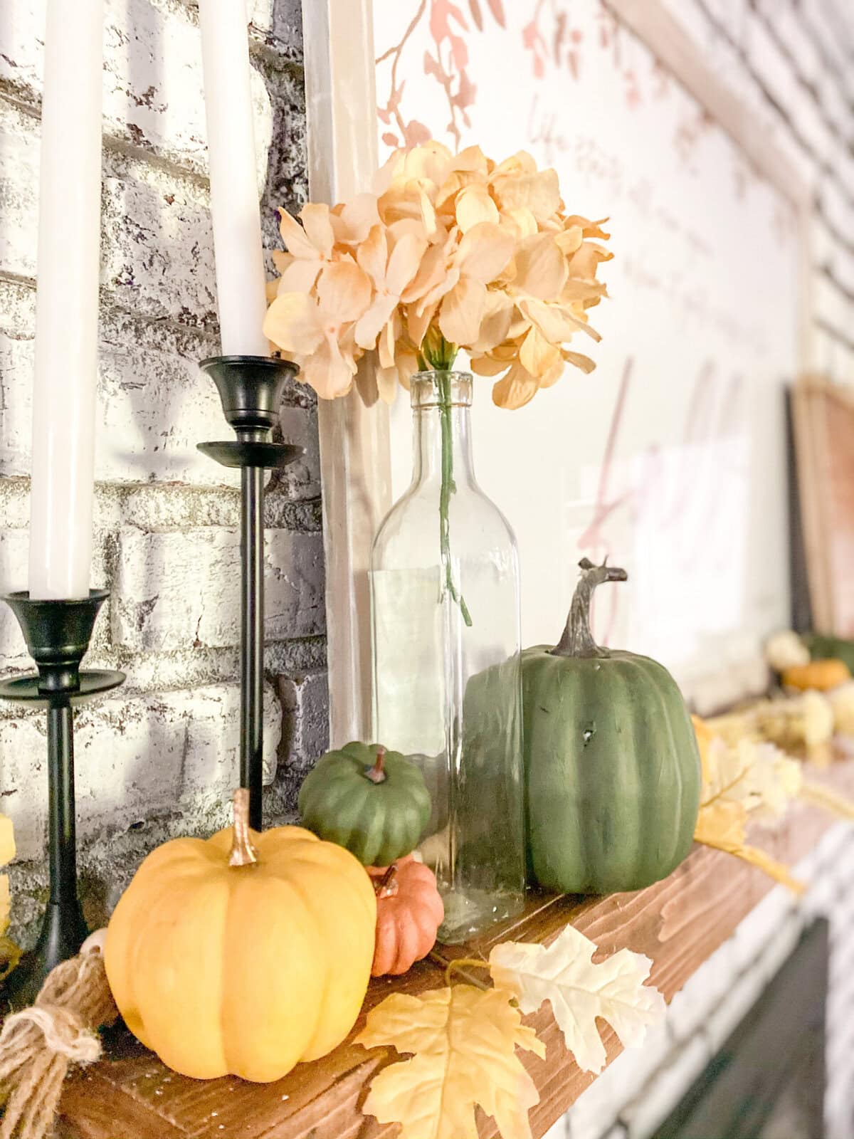 Vase with peach hydrangeas and decorative pumpkins on a rustic wooden mantel with candles, autumn leaves, and a brick wall, creating a warm, cozy fall decor scene for a lifestyle blog or home inspiration.