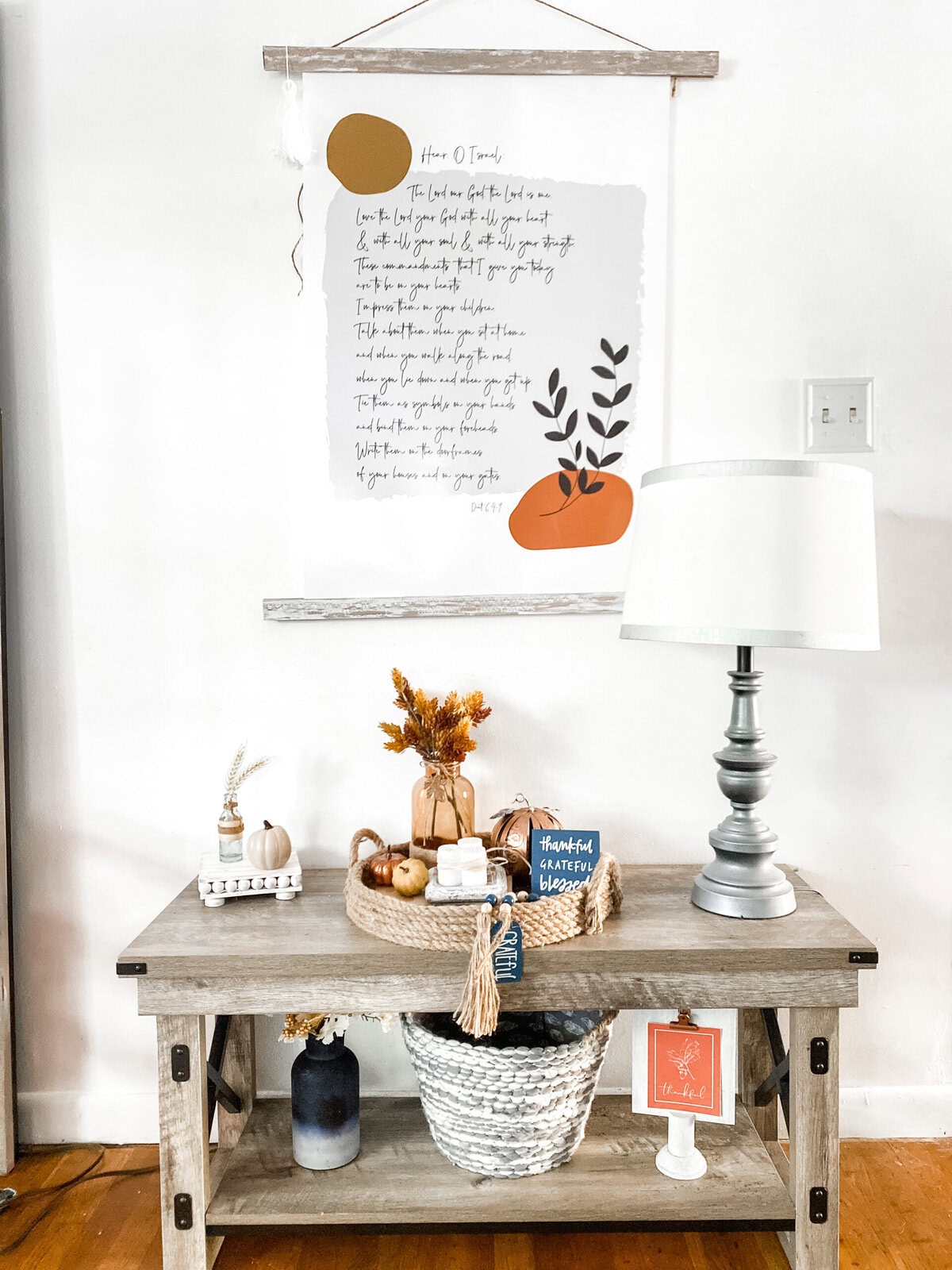 Rustic wooden console table decorated with fall-inspired accents, greenery, pumpkins, and gratitude-themed decor, topped with a white table lamp against a neutral wall.
