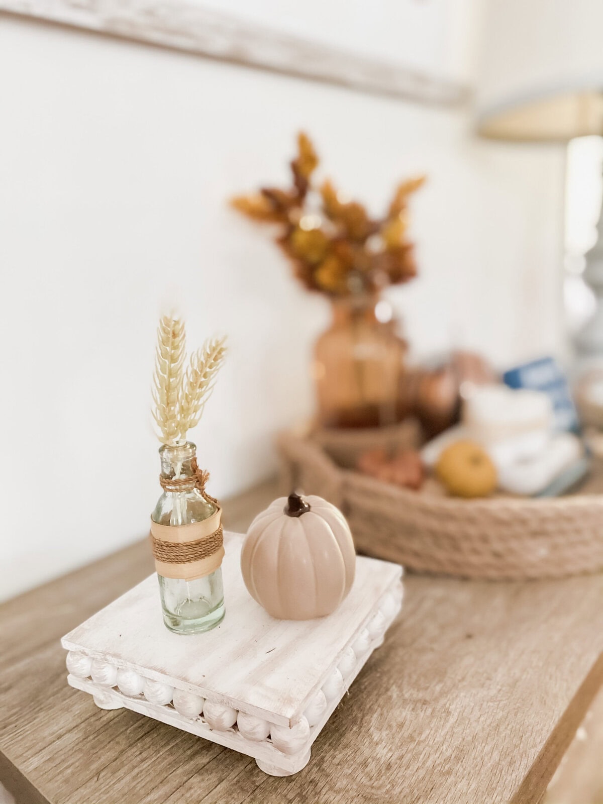 Warm neutral fall decor featuring a miniature pumpkin, dried wheat in a small glass bottle, and soft earthy tones creating cozy autumn ambiance.