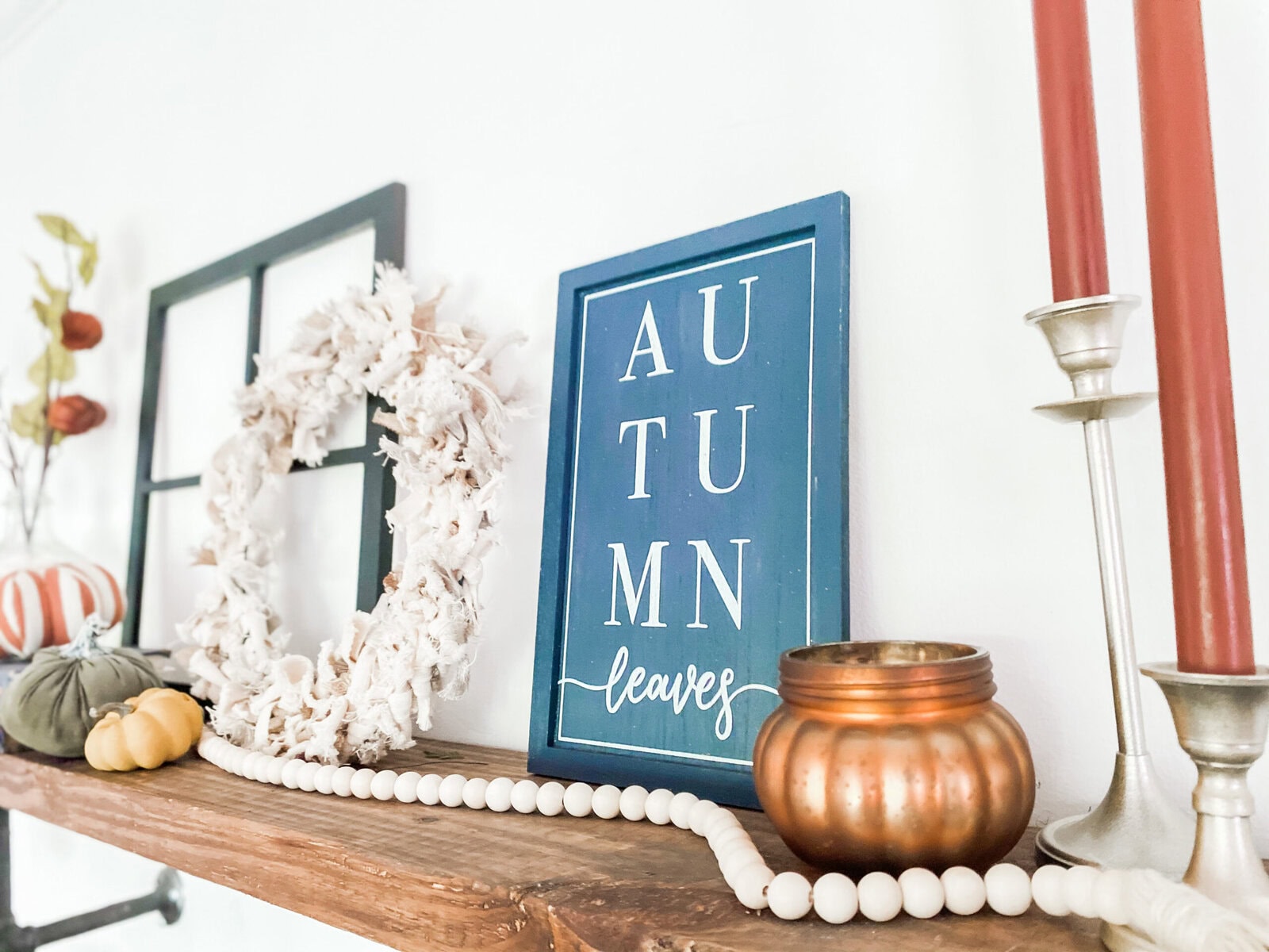 Bright fall-themed home decor display featuring pumpkins, candle holders, and a framed wall art with the phrase "Autumn leaves" on a wooden shelf. Perfect for cozy seasonal home accents.
