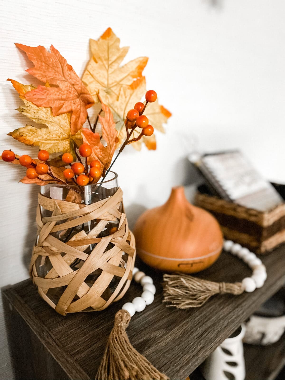 Vase with fall-themed artificial leaves and orange berries, decorative pumpkin, and wooden beads with tassels on a dark wooden surface for autumn home decor.