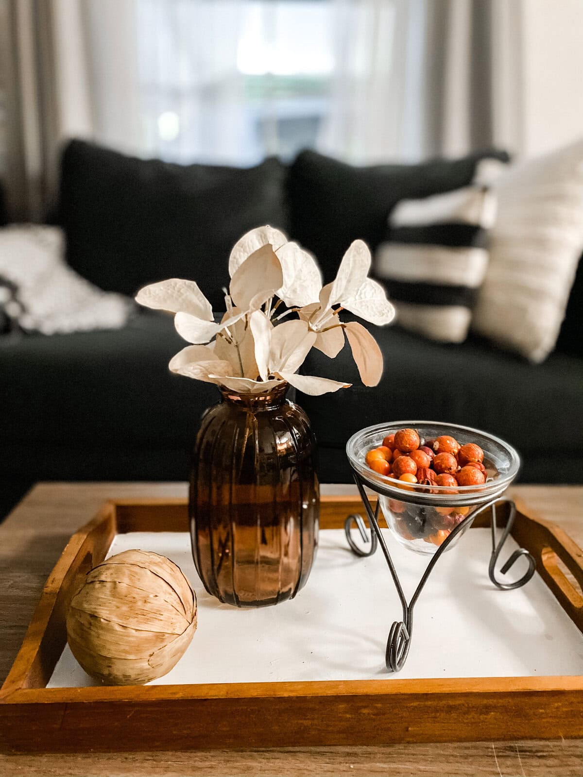 Elegant autumn-inspired home decor arrangement with a brown glass vase, dried leaves, a wooden decorative ball, and a tray featuring hazelnuts, creating a cozy, stylish living space atmosphere.