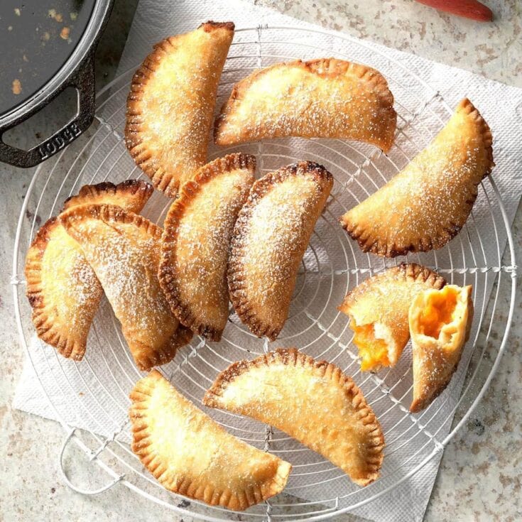 Golden fried empanadas with a dusting of powdered sugar on a wire cooling rack, showing a flaky, crispy exterior and one with a glimpse of sweet filling inside, perfect for dessert or snack.