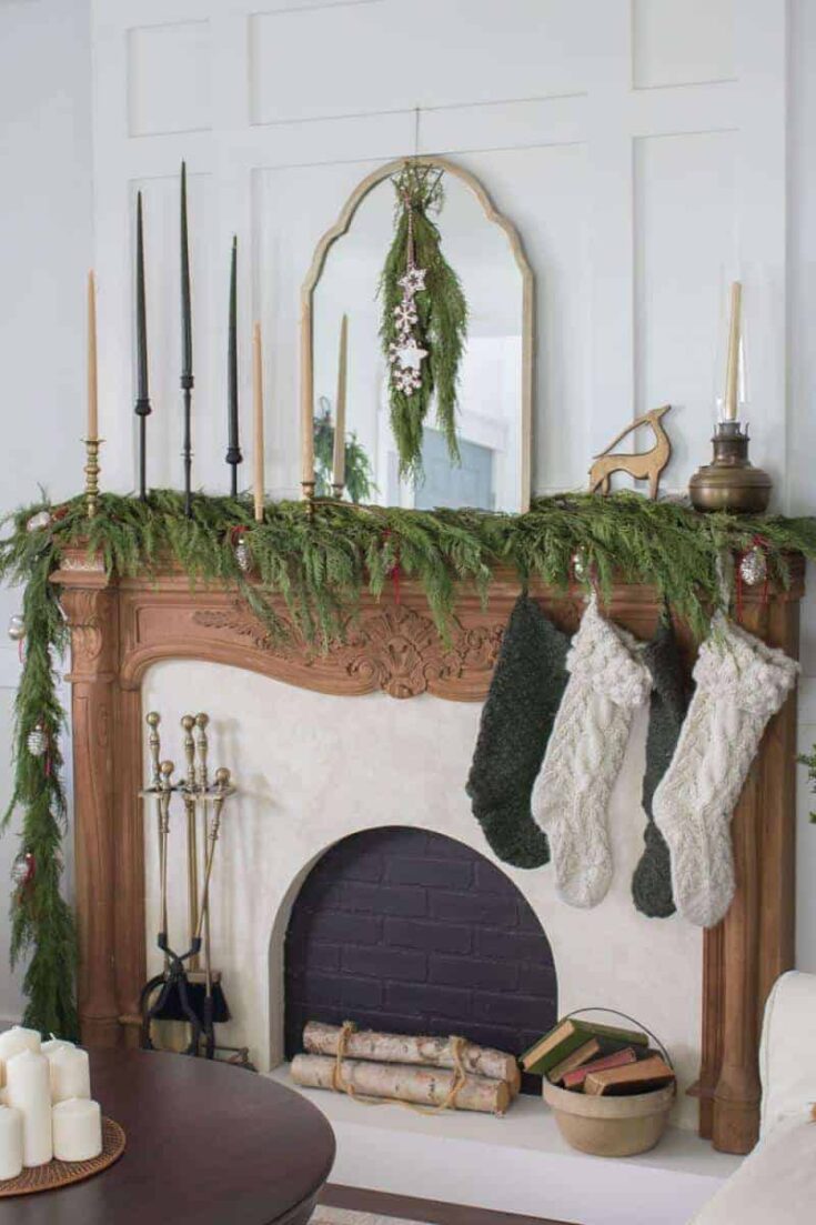 Festive Christmas fireplace decorated with greenery, candles, stockings, and a mirror, creating a cozy holiday setting with rustic charm and seasonal decor.