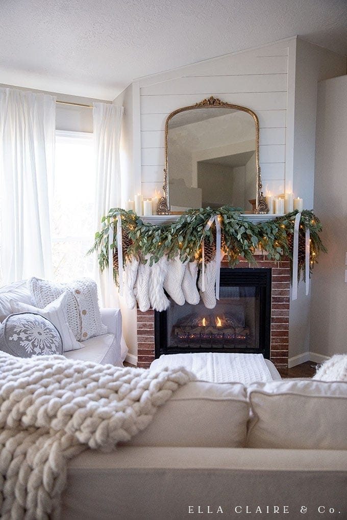 Cozy living room fireplace with holiday garland, stockings, candles, and a mirror, enhancing festive home decor for Christmas or winter seasons.