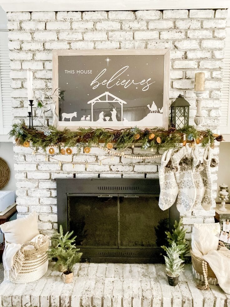 Festive Christmas mantle with greenery, pinecones, and holiday decor featuring a sign that reads "This house believes" against a white brick fireplace.