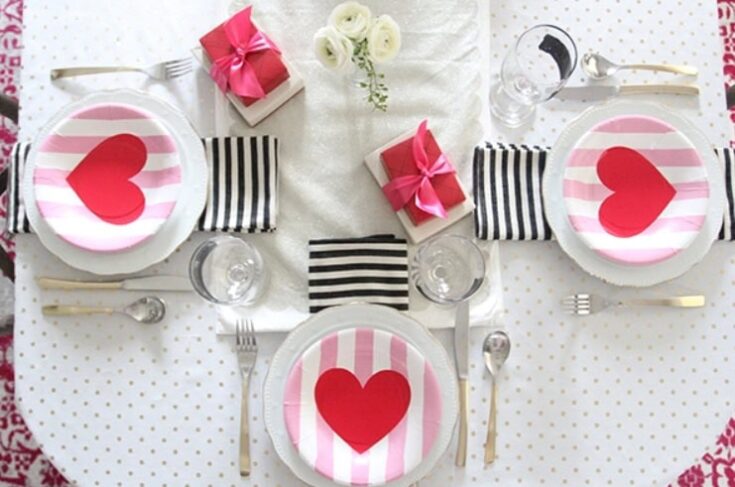 A festive Valentine's Day table setup featuring pink and red heart-themed plates, elegant cutlery, wrapped gifts, and floral accents arranged for a romantic celebration.