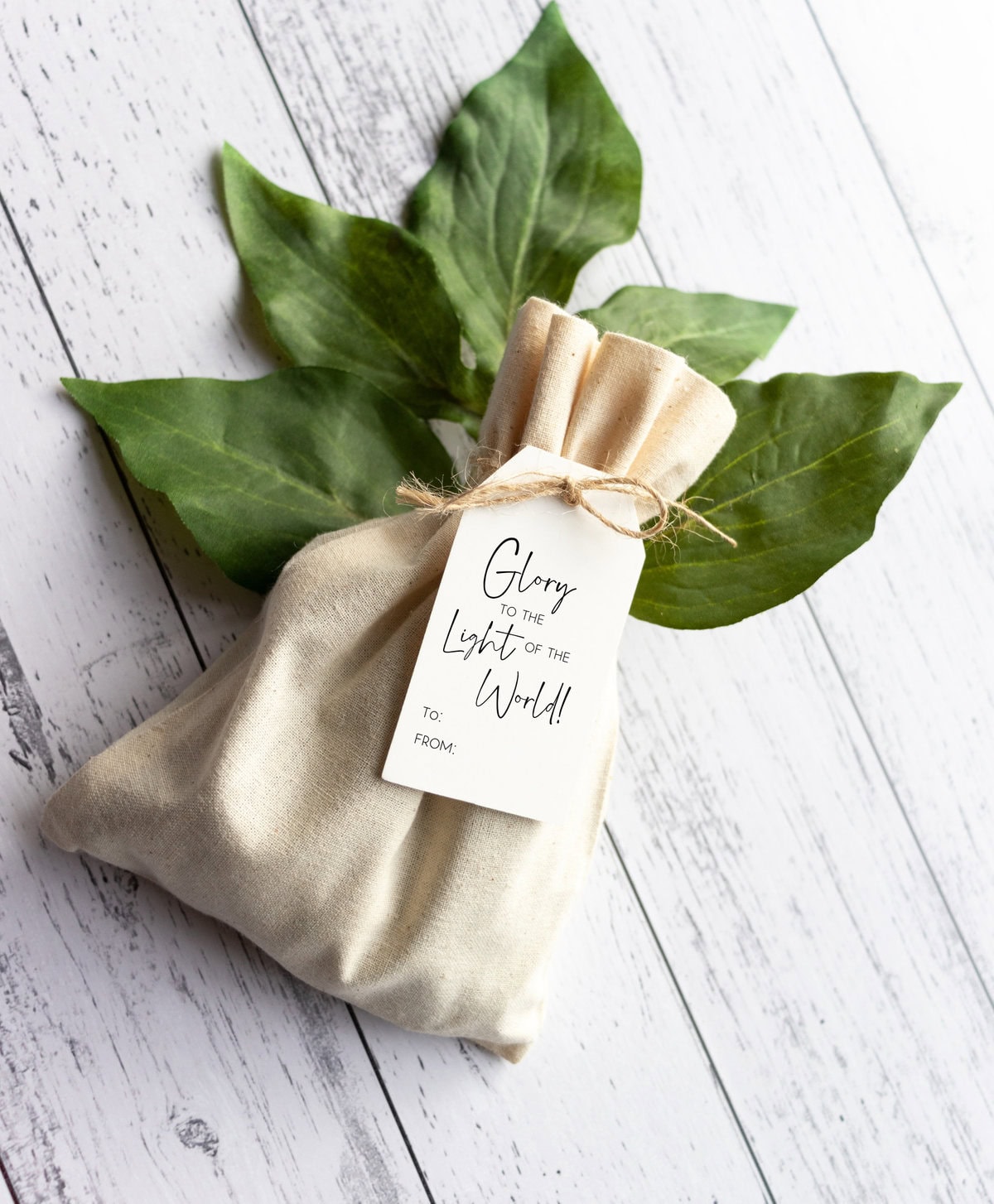 Gentle white fabric gift bag with a botanical green foliage accent, tied with twine and a heartfelt "Glory to the Light of the World!" gift tag for Christmas or special occasion celebrations.
