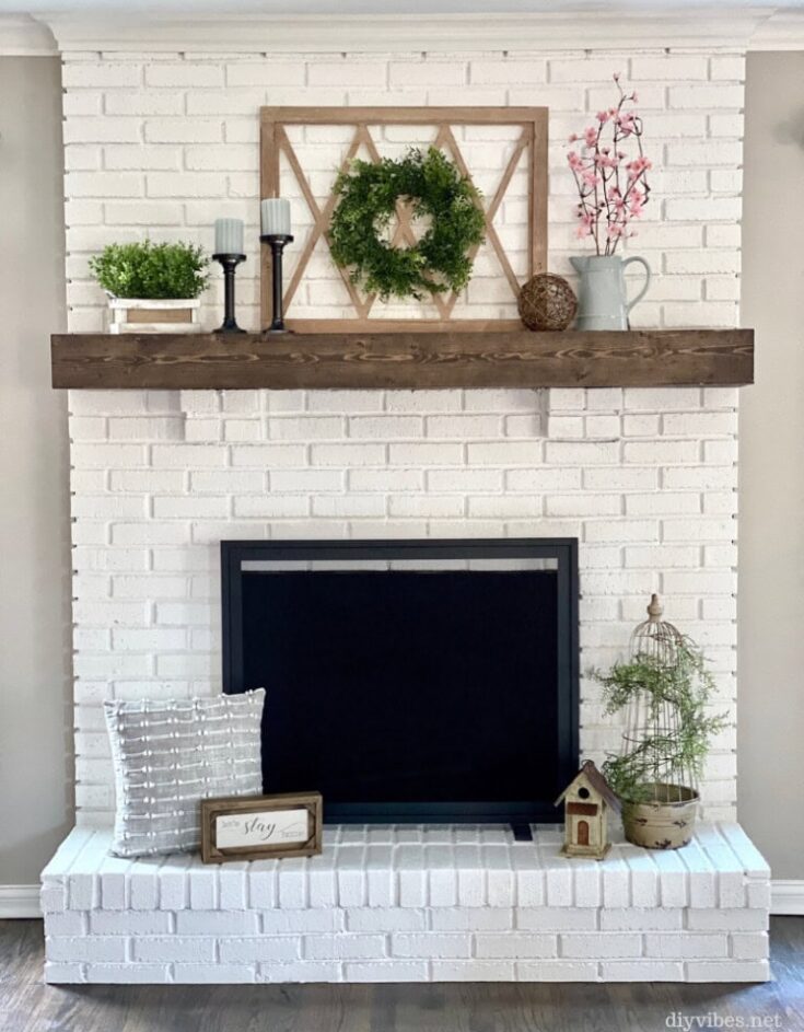 Vintage white brick fireplace with wooden mantel decorated with greenery, candles, and pink blossoms, creating a cozy and charming focal point for home decor.