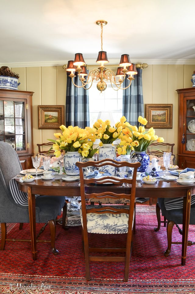 Elegant dining room with antique wooden table, blue and white porcelain vases filled with yellow tulips, classic chandelier, framed artwork, and vintage furniture for a charming home decor look.