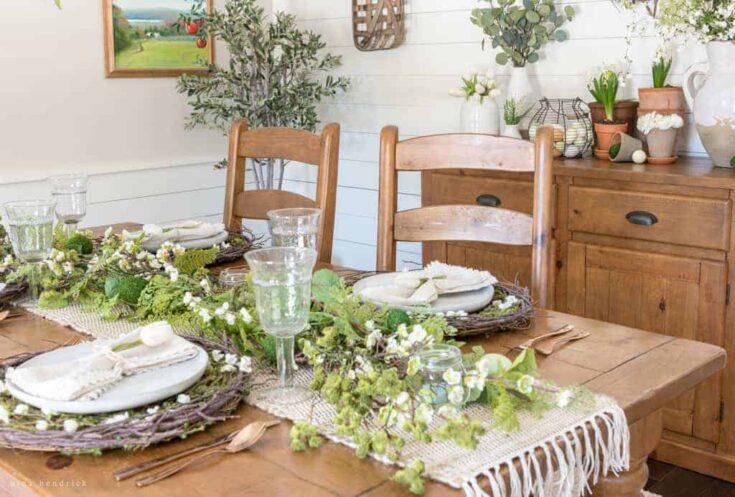 Wooden dining table with floral spring centerpiece, set for a meal with white plates, cloth napkins, and glassware, in a rustic farmhouse with natural wood furniture and green plants.