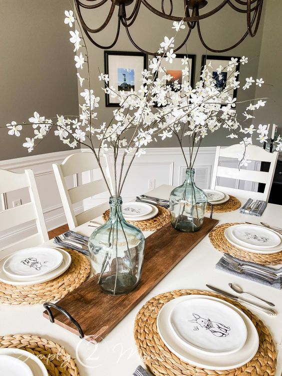 Linen table setting with white plates featuring bunny illustrations, woven placemats, glass vases with white blossoms, and a wooden centerpiece, in a cozy dining room with framed artwork and a chandelier, perfect for spring or Easter celebrations.