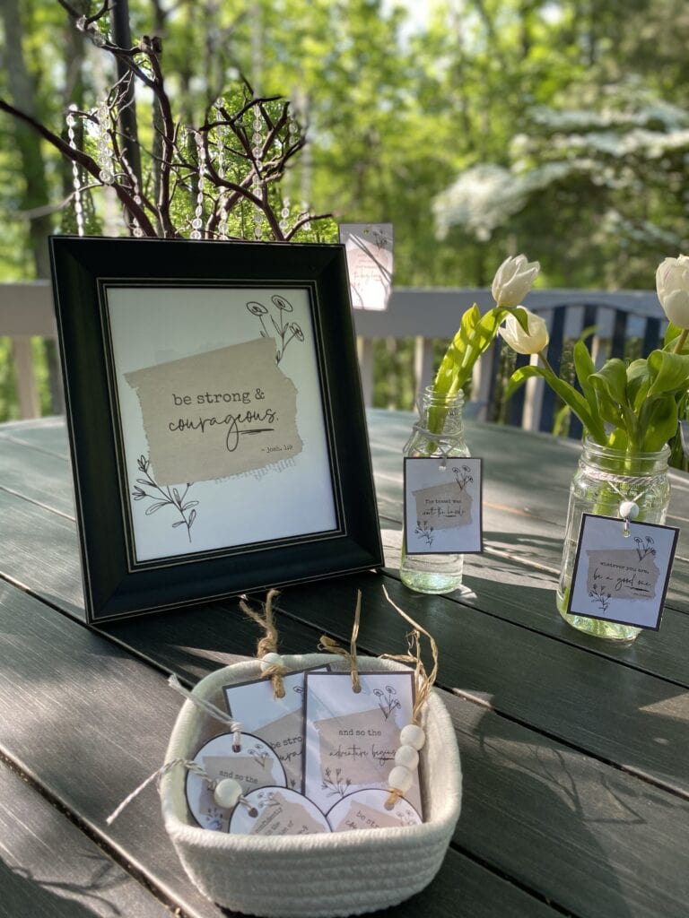 Vase with white tulips and inspirational quotes displayed on a black picture frame and jar tags, set outdoors on a weathered table with a lush green background.