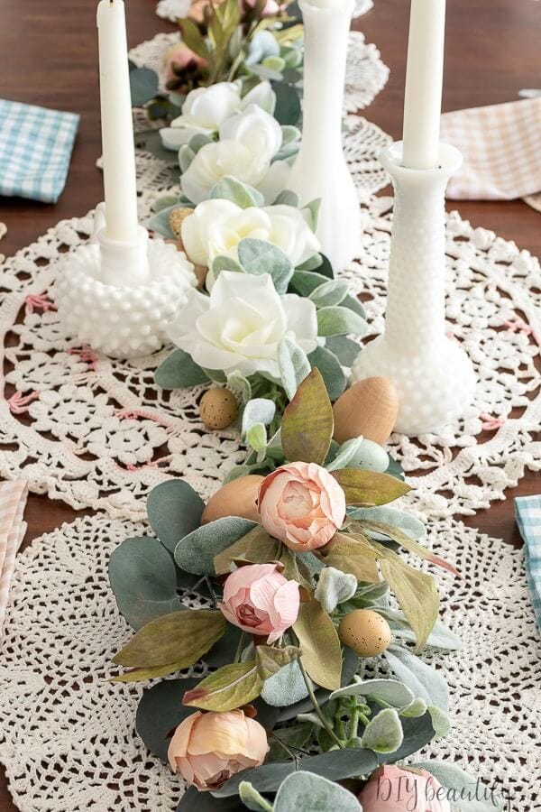 Soft white candles with textured bases, elegant white flowers, and muted faux eggs on a delicate lace table runner create a charming and romantic centerpiece for spring or Easter celebrations.