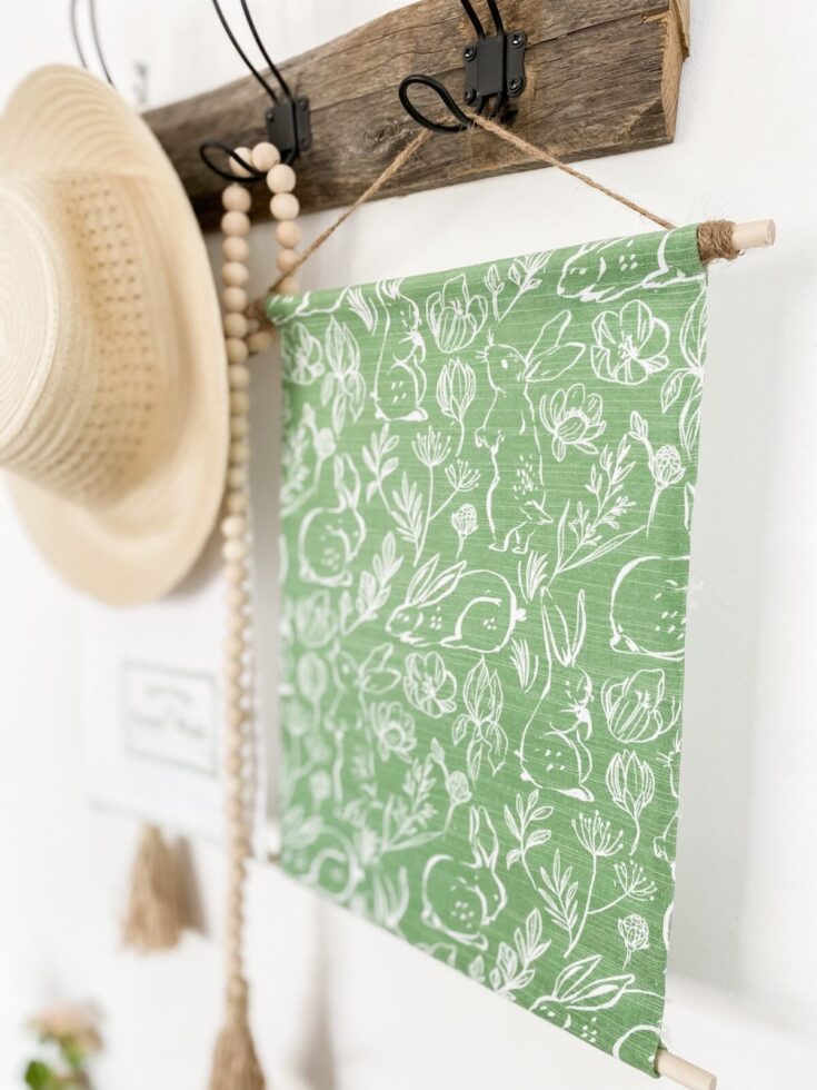 Vibrant green fabric wall art with white bunny and floral illustrations, hanging on a wall-mounted wooden and black metal hook system, adding a charming and artistic touch to home decor.