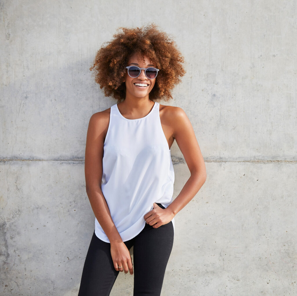 Vibrant woman with curly hair in sunglasses and white tank top smiling outdoors against a concrete wall, showcasing confidence and joyful style.