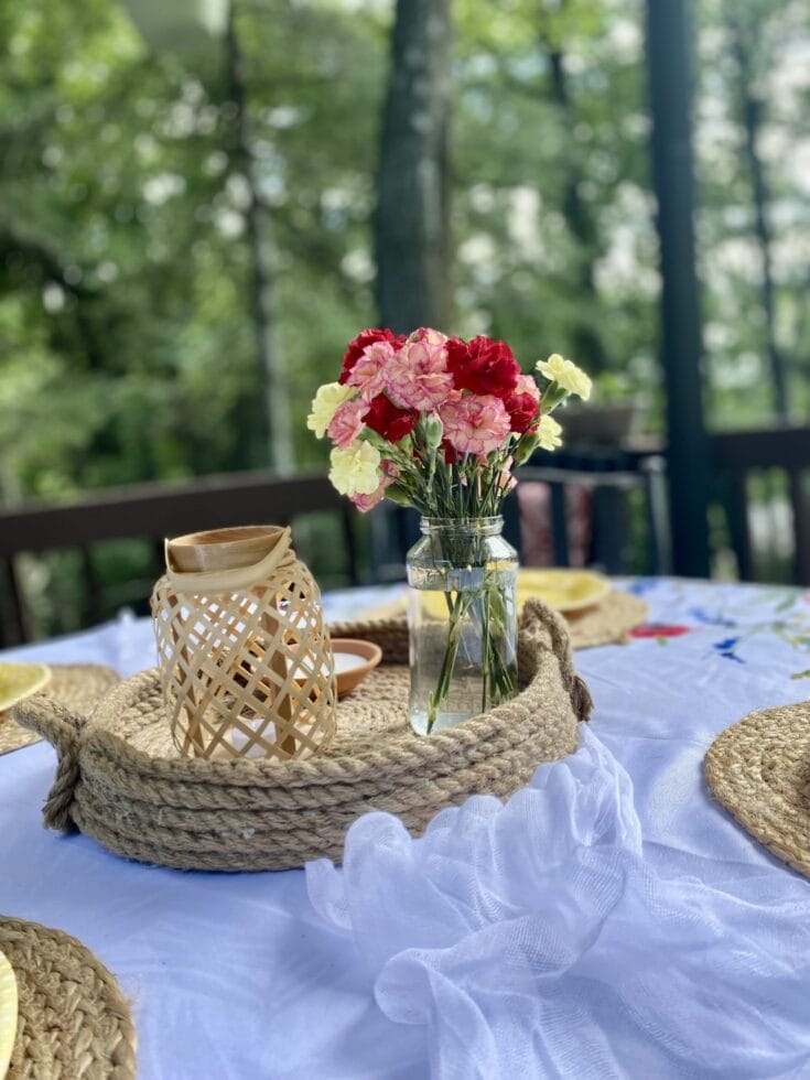Vase of pink and red carnations on a white outdoor table with woven placemats, surrounded by greenery for a charming, nature-inspired setting.