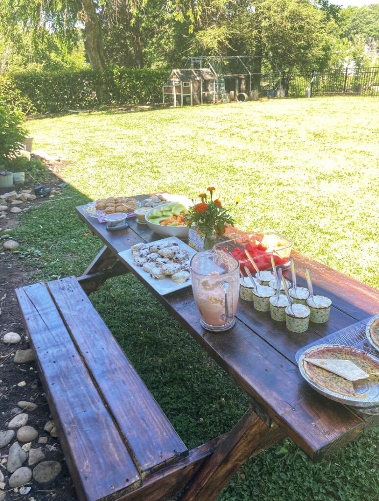 Fresh outdoor picnic table with homemade baked goods, fresh fruits, and desserts set in a lush backyard garden for a family gathering or celebration.