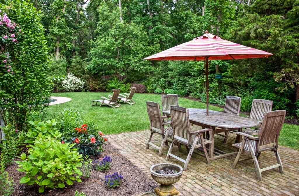 Rustic outdoor dining area with wooden table, chairs, and large red striped umbrella in lush green garden. Perfect for relaxing and entertaining in a natural backyard setting.
