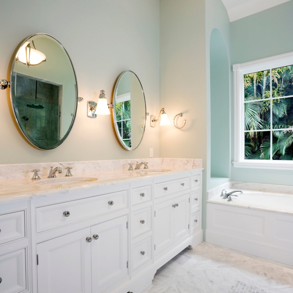 Elegant light blue bathroom featuring a vintage clawfoot bathtub, white cabinetry with marble countertops, and large windows with scenic views, creating a serene and luxurious space.
