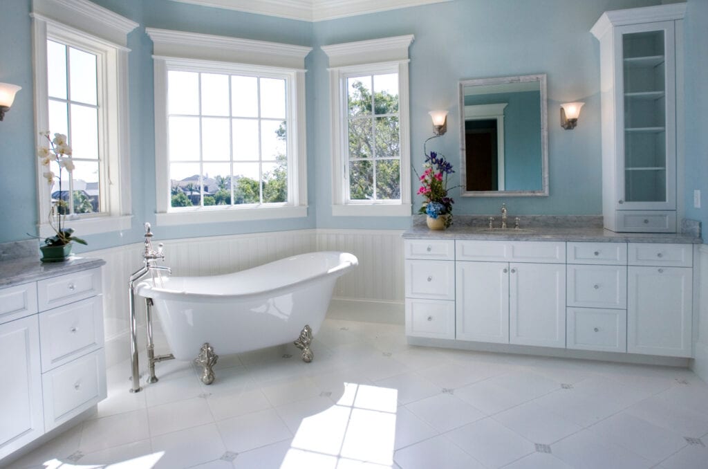 Elegant light blue bathroom featuring a vintage clawfoot bathtub, white cabinetry with marble countertops, and large windows with scenic views, creating a serene and luxurious space.