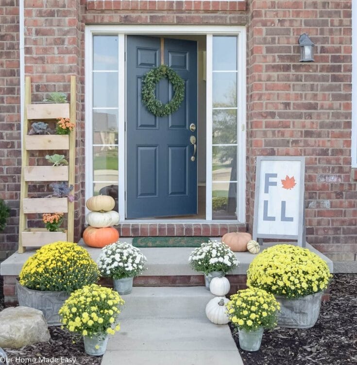 Warm fall front door decor with pumpkins, chrysanthemums, and a green wreath, complemented by a wooden plant ladder and seasonal "FALL" sign, creating a cozy autumn porch aesthetic.
